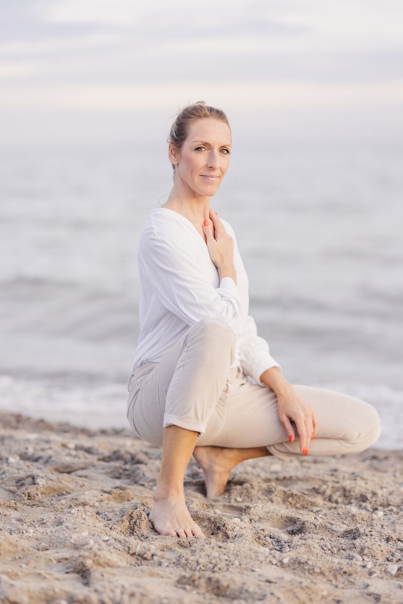 Renée hockt barfuß am Strand, eine Hand auf dem Herzen, und schaut Dich an